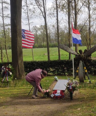 Een oude dame bukt om bloemen te leggen bij een steen met een plakette van slachtoffers in de Tweede Wereldoorlog in Herkenbosch. Een jongeman van een jaar of 18 kijkt toe.Op de achtergrond bomen, een grasveld en de wapperende vlaggen van Nederland, Engeland en de Verenigde Staten.